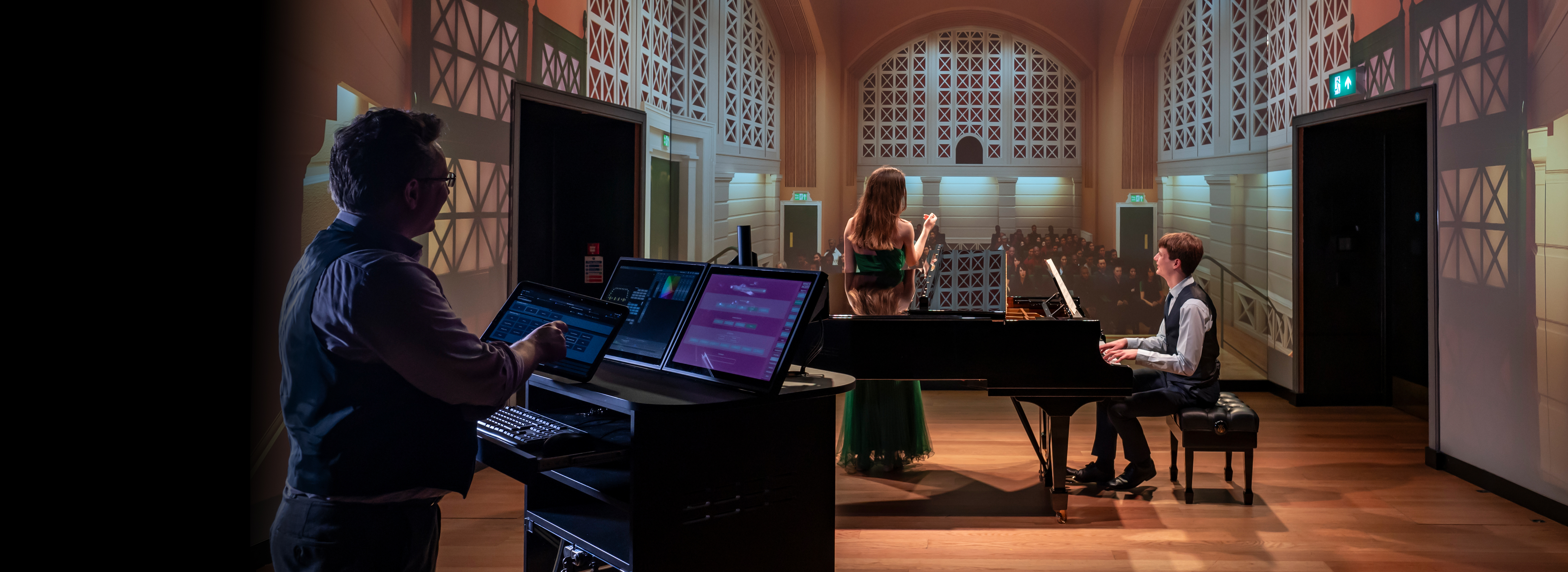 A male and female student, wearing formal attire, singing and playing the piano, in front of a performance simulator.